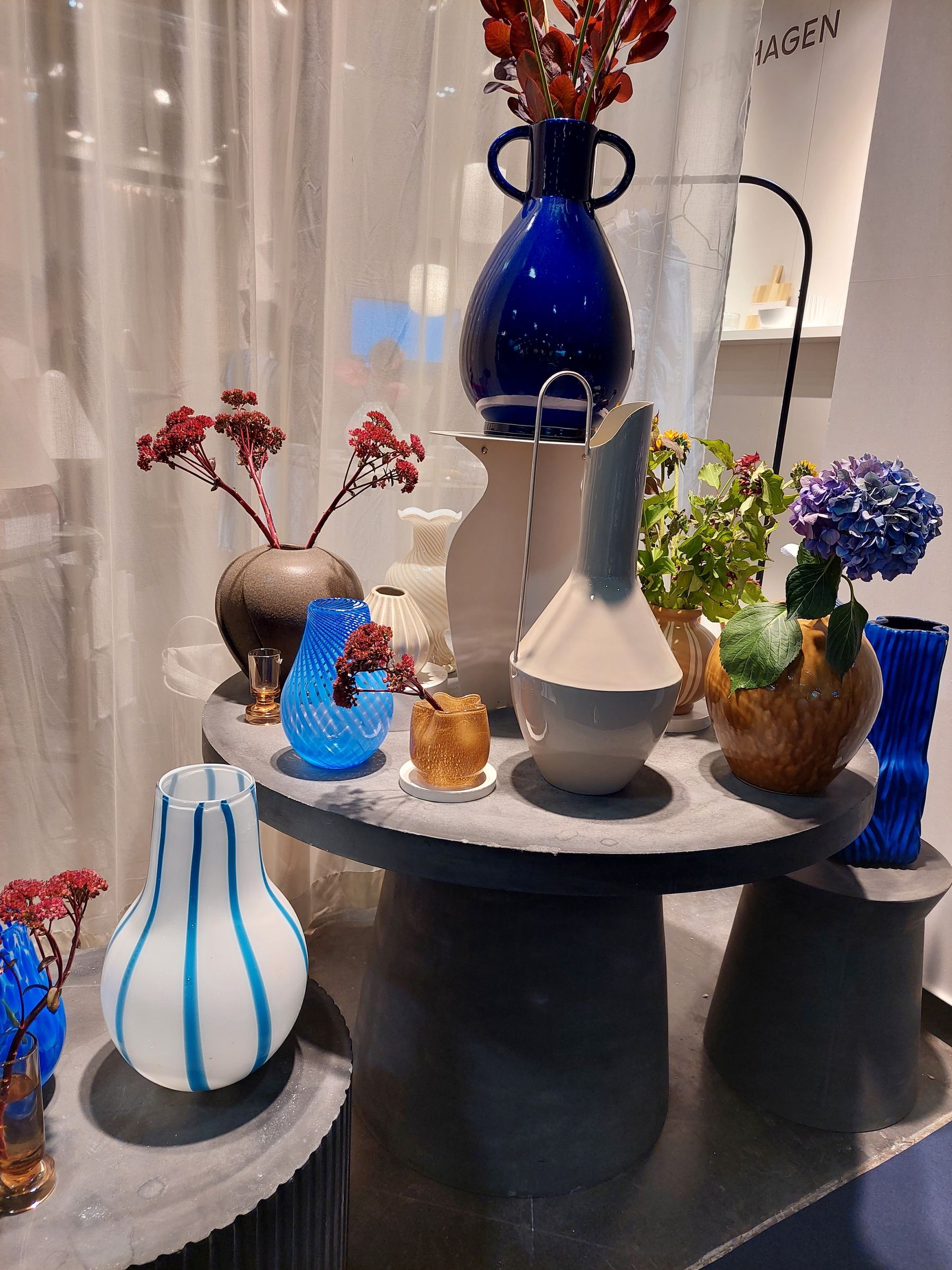 Arrangement aus Glasvasen in Blitzblau und Creme mit Blüten, auf verschieden hohen Beistelltischen