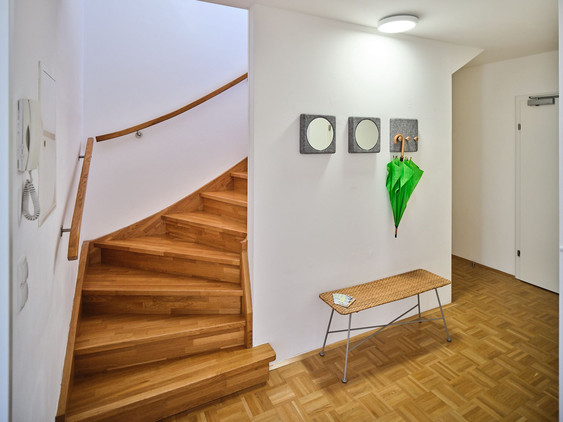 Vorzimmer mit Parkettboden und Stiegenaufgang in Holz, Sitzbank Metall mit Korbsitzfläche und 3teilige quadratische Filzgarderobe 20x20 mit Spiegel und Haken, darauf ein grüner Stockschirm