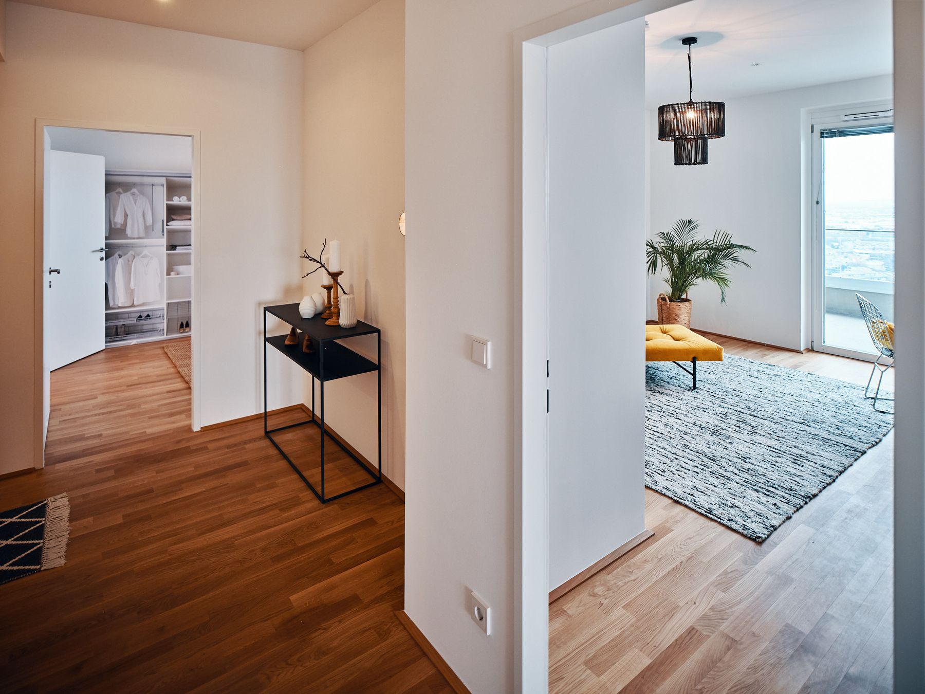 Eingang in L-Form mit Holzboden und schwarzer Metallkonsole und Blick ins Schlafzimmer sowie in ein Arbeitszimmer mit senfgelbem Daybed, schwarzer Rattanleuchte und weiß-schwarzem Wollteppich und einer Palme im Seegras-Übertopf in der Ecke