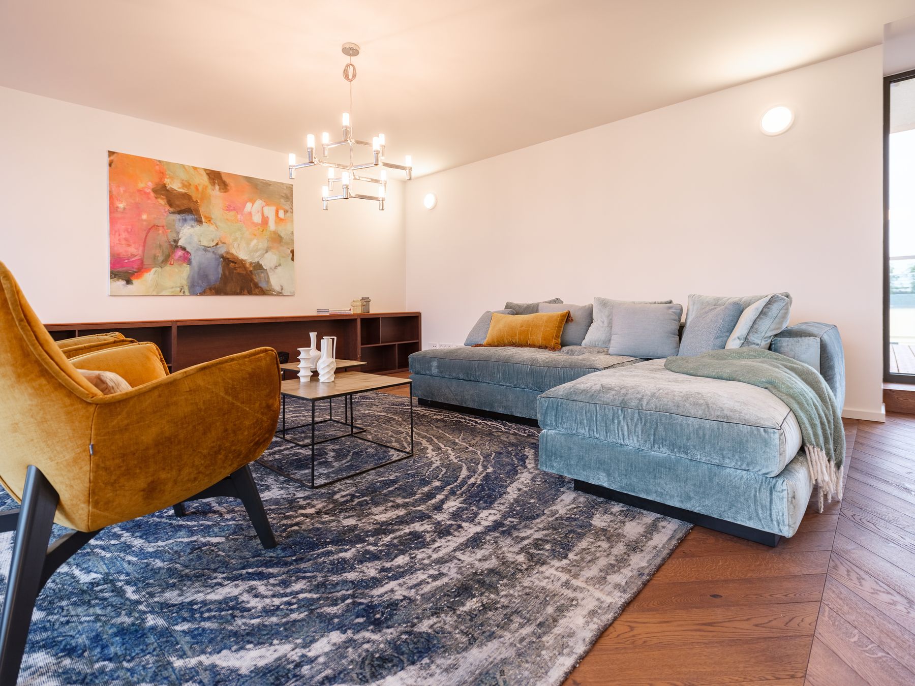 großer Wohnraum mit mintgrünem Samtsofa goldig-schwarzem Couchtisch-Set großen gelben Samt-Loungestühlen Vintageteppich in blau mit Kreisen sowie Design-Chrom-Luster 12-armig mit matten Glaszylindern und abstaktem Bild in kräftigem Pink-Gelb-und Braunton