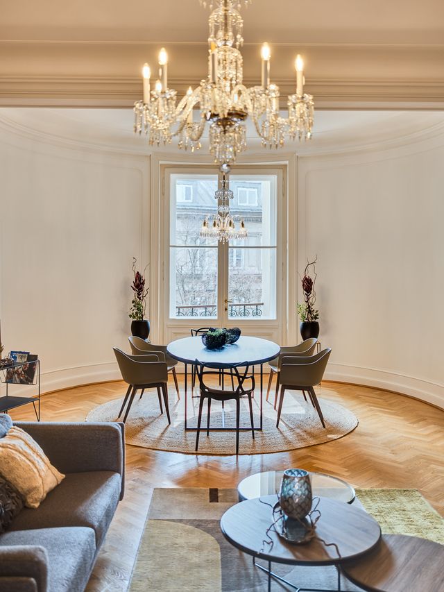 Wohnzimmer mit Luster, grauem Sofa, Couchtisch-Set aus Metall mit Holzplatte, Wollteppich in grau-gelb-Tönen, und im runden Erker massiver Esstisch aus Holz mit schwarzem Metallgestell, Design-Stühle mit Holzbeinen und salbeifarbenen Kunststoffschalen
