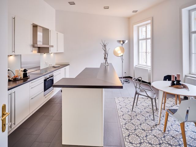 Wohnküche weiß mit schwarzer Arbeitsplatte, rundem Tisch in weiß mit 4 Stühlen in grau, Teppich mit Blumenmuster und große Stehleuchte mit 2 halbrunden silbernen Schalen