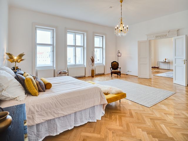 Schlafzimmer mit Boxspringbett, Wollteppich, schwarzen Nachttischen, altem Lesesessel in blau-gelb, gelber Sitzbank und Stehleuchte aus Glas sowie traditionellem Luster