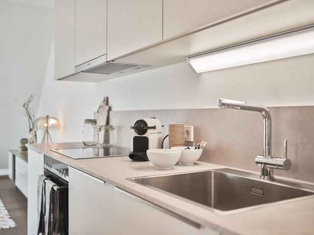 Einbauküche in weiß mit grauer Platte inkl Oberschränken, Nespressomaschine und Becher, Geschirrtücher an der Herdstange, Blick ins Wohnzimmer Richtung Lowboard