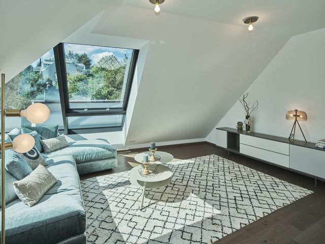 Dachgeschoß-Wohnraum mit 2 Deckenspots, goldene Stehleuchte mit Bulbs im Vordergrund, L-förmiges Samtsofa in mintgrün, Teppich Baumwolle creme mit schwarzen Rauten, 3-Ebenen-Couchtisch Metall in taupe, Lowboard in weiß/grau mit Laden, Tischleuchte gold