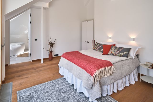 Schlafzimmer Bett mit grauer Bettdecke und rot-grauen Accessoires, grauer Nachttisch, rote Bodenvase und grauem Teppich sowie Wandleuchten in Würfelform