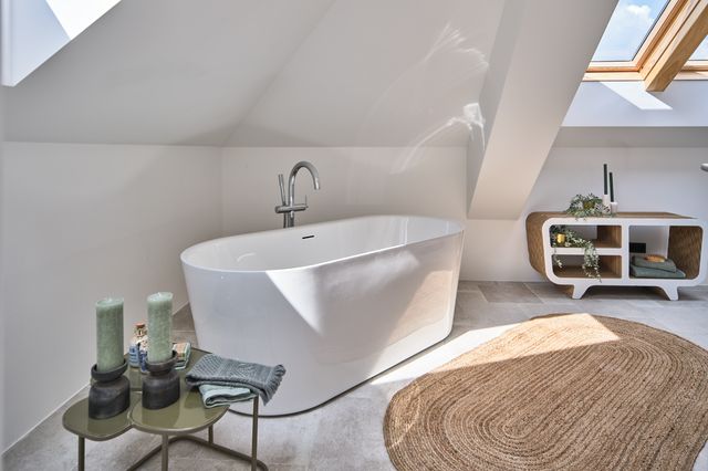 Bad mit grauem Steinboden, Sisalteppich, freistehender weißer Wanne, Metall-Beistelltischen, dunklen Holzkerzenleuchtern und salbeifarbenen Handtüchern sowie Sideboard mit salbeifarbener Deko