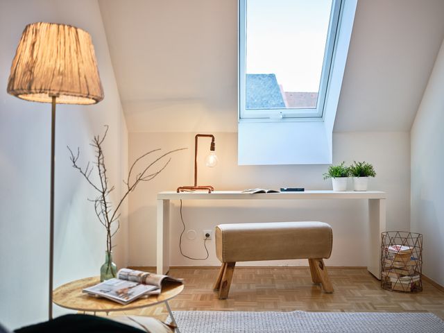 Mini-HomeOffice: Blick vom Sessel auf Stehleuchte mit Stoffschirm, Beistelltischchen aus Sperrholz, weiße Hochglanz-Konsole mit Sitzbank wie eine alte Turnbank, Tischleuchte aus Kupferrohren mit Glühbirne, Gitterkorb Kupfer mit Büchern, grüne Glasvase