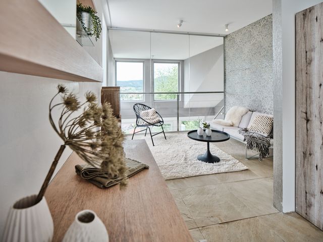 Galerie symbolisiert als Gästezimmer mit Sofa in beige, rundem Couchtisch in schwarz und Kuschelteppich in creme, dazu Holzregale mit Vasen und Blüten und Stehleuchte mit Kugeln in creme