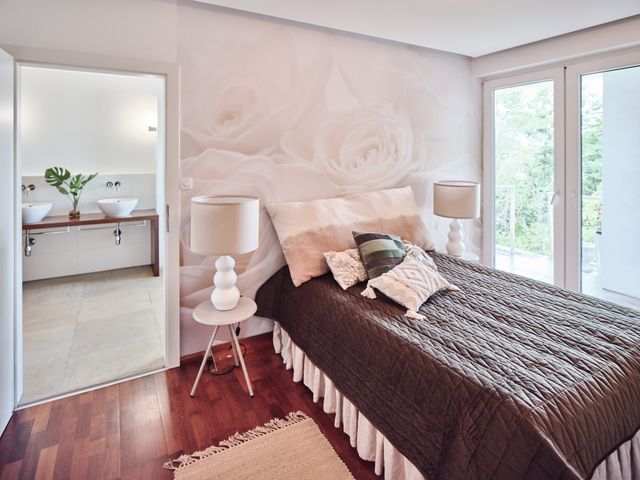 Schlafzimmer mit grauer Tagesdecke und hellen Kissen, Beistelltisch und großen Tischleuchten mit hellen Schirmen, angrenzend das Badezimmer mit Doppelwaschtisch aus Holz und weißen Aufsatzbecken