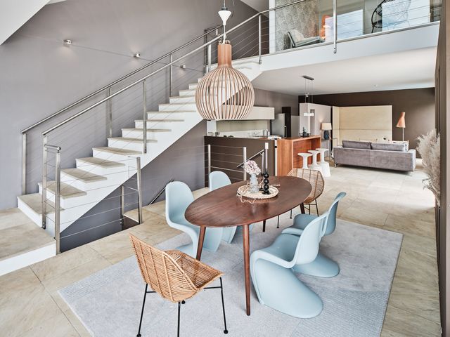 Essplatz neben der Stiege mit grauer Wand, ovaler Holztisch und verschiedene Stühle in Rattan, Pantone Chairs in eisblau und eine Secto Leuchte aus Holz mit weichem Teppich in graublau