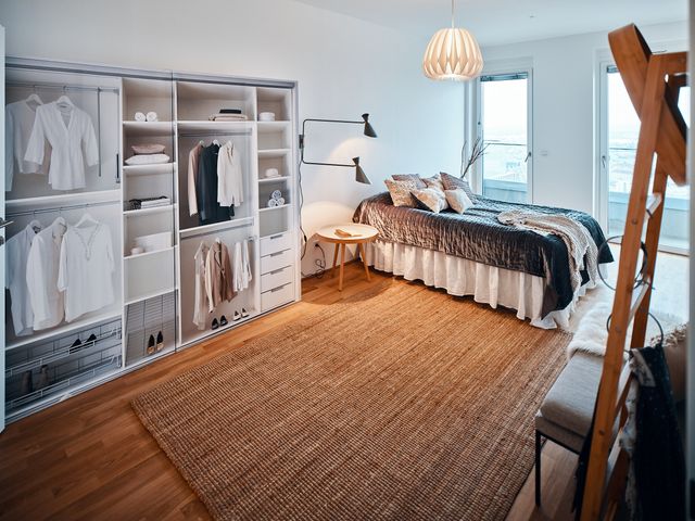 Schlafzimmer mit Dummy-Schrankwand, Doppelbett mit grauem glänzendem Überwurf, weißer Papierfaltenleuchte, Wandleuchte zweiarmig grün als Leselicht und kleinem Beistelltisch aus Holz, Sitzbank und Stehgarderobe wie Holzleiter und Sisalteppich