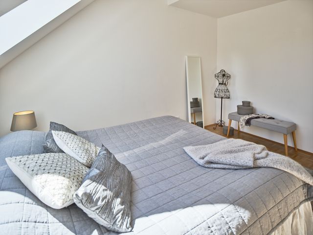 Bett mit grauer Bettdecke und unterschiedlichen Zierkissen in Grautönen und Strickplaid, graue Tischlampe, Sitzbank grau mit Holzbeinen und Dekoschachteln, Standspiegel weiss, Kleiderpuppe aus Metall