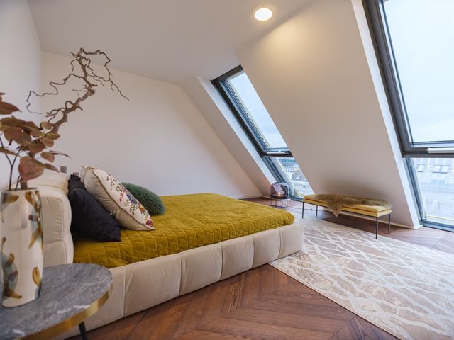 Doppelbett mit wuchtigem Stoffbezug in creme und senfgelber Überdecke mit bunten Kissen, davor Sidetable in Marmor und Vase mit Zweigen, dahinter senfgelbe Sitzbank auf cremefarbenem Teppich und Bodenleuchte mit halbrundem getöntem Glasschirm und Messing